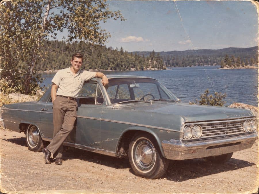 A faded, creased 1960s snapshot of a young man leaning on his car beside a lake